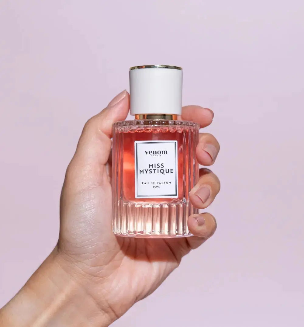 Hand holding a bottle of 'Miss Mystique' perfume against a light pink background.
