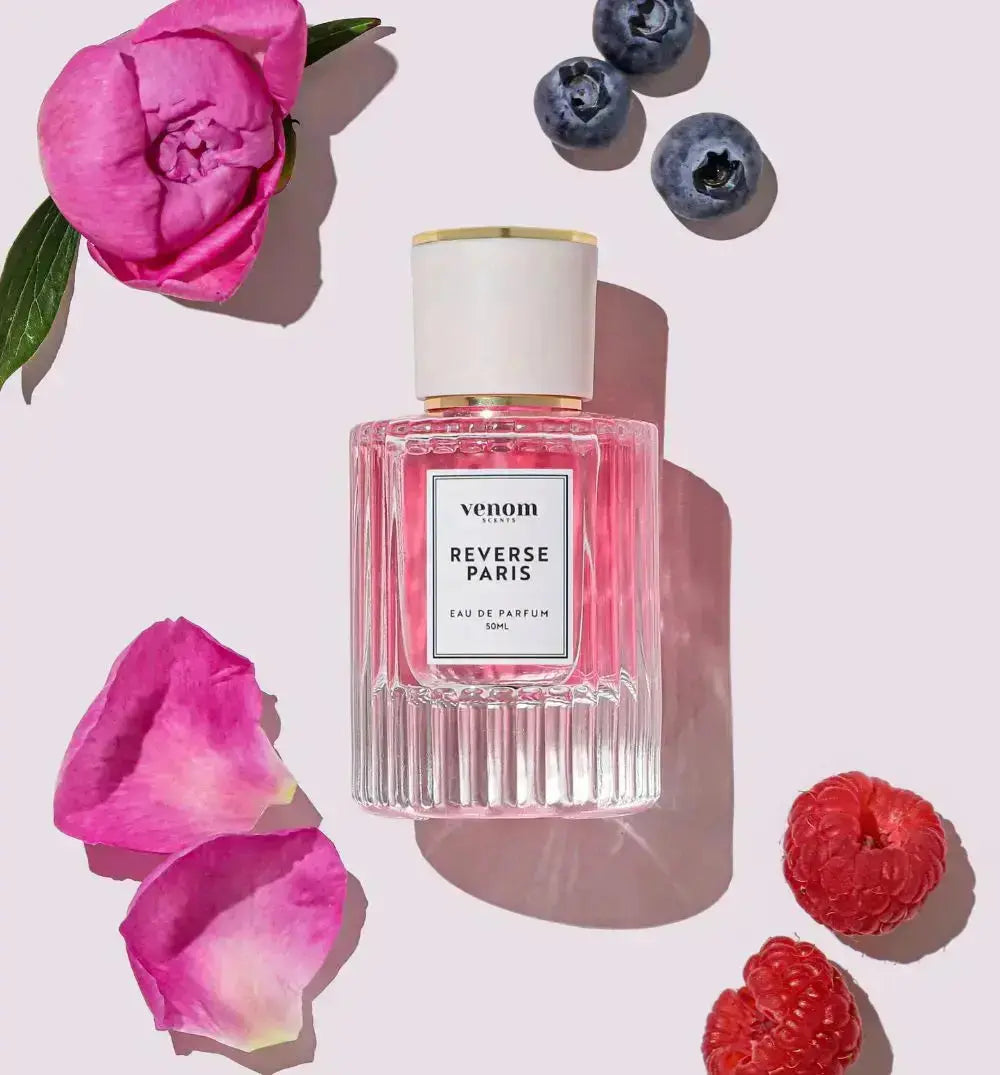 Perfume bottle with pink flower petals, blueberries, and raspberries on a light background.